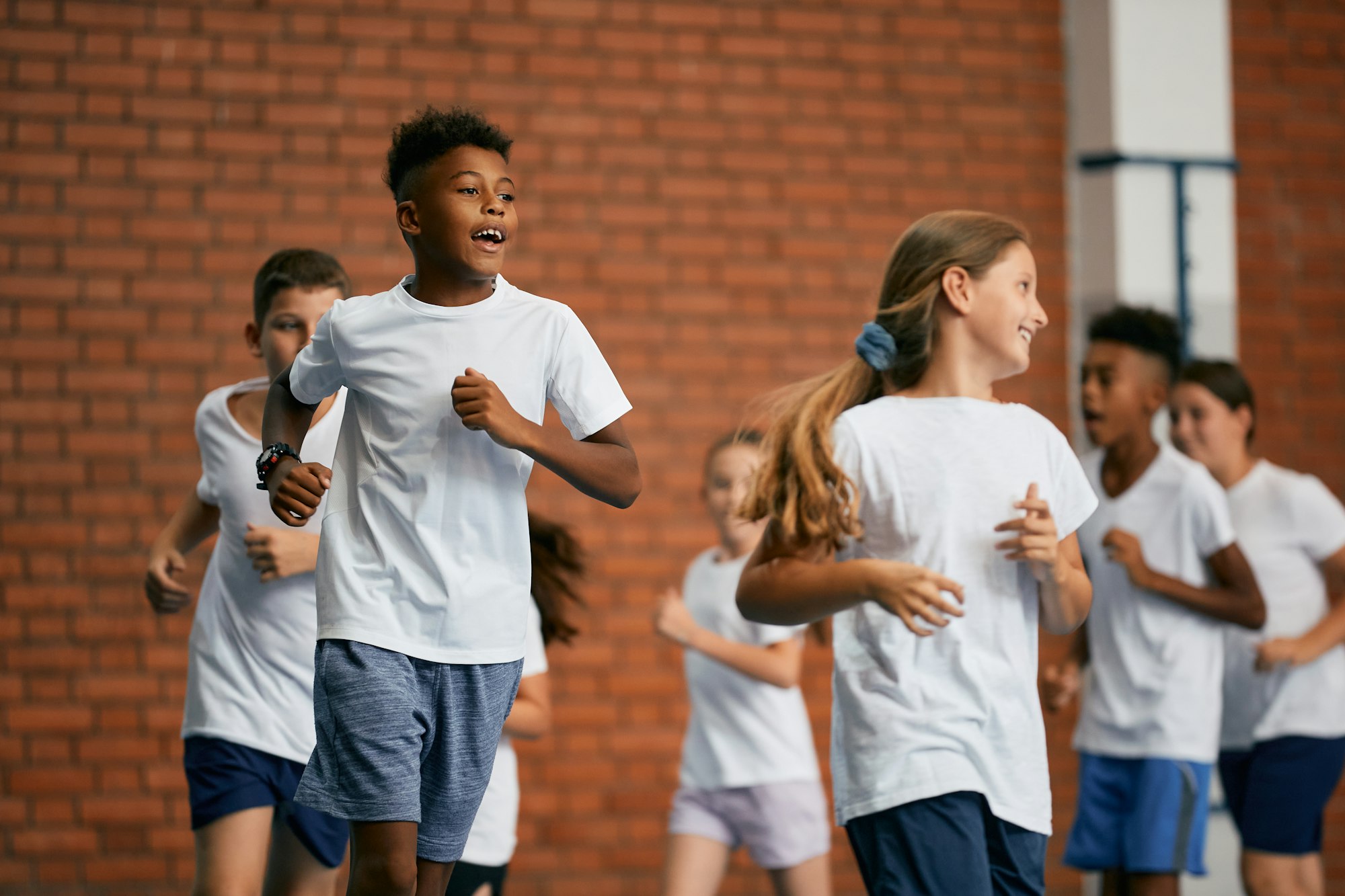 Black elementary student and his friends running on physical education class at school gym.